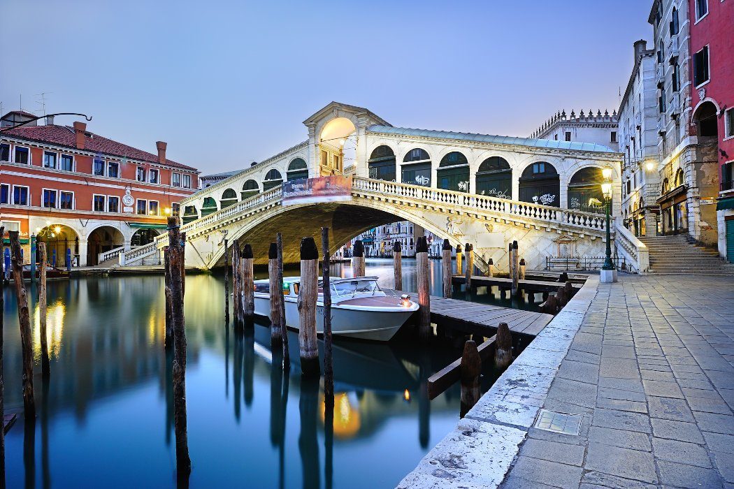 Photo of Rialto Bridge in Venice at dawn before the onslaught of tourists in the morning. The streets are completely deserted, and the tranquility is reflected in the canal, with nary a ripple disturbing its serene beauty. This is the perfect place to stay for you trip to Venice if you have the budget.